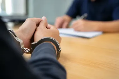 Skute kajdankami dłonie osoby siedzącej przy drewnianym stole naprzeciwko funkcjonariusza piszącego w notatniku.